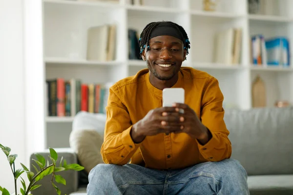 African American using a smartphone, browsing the Internet, sitting on the couch at home. Black man texting on cell phone or using new app on cell phone indoors. High quality photo