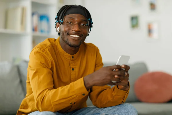 Smiling man using phone on internet while sitting on sofa at home. Cheerful mobile phone user testing new smartphone app, posing, looking at camera. High quality photo