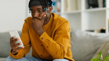 Sad young black american guy sitting on the couch, looking at the phone,. High quality photo