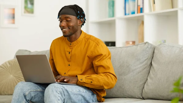 Smiling black man using computer working or studying while sitting on sofa at home. Free copy space. High quality photo