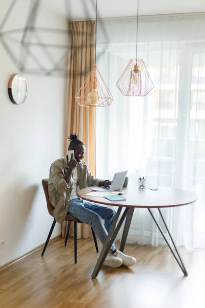 Young black man speaks on the phone, uses a laptop at a table in a home office. Cool african american man working remotely, freelancing online. Modern business concept. High quality photo