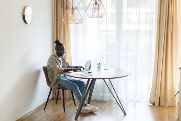 Happy young black man using laptop computer for online work at table in home office. Cool African American guy having remote job, freelancing on web. Modern business concept. High quality photo