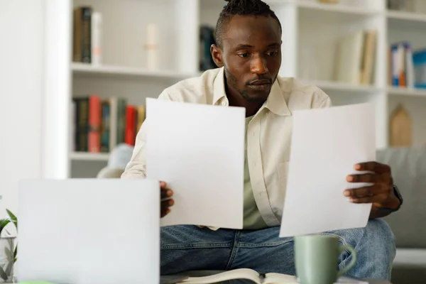 Young african man sitting at table with modern laptop in home office, reading paper mail, holding document or bills, thoughtful guy got bad news, notice of debt. High quality photo