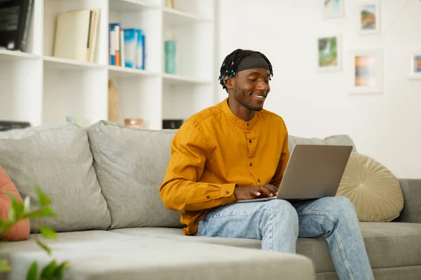 Black african man in a shirt with a laptop sits on a sofa in the living room, free copy space. High quality photo