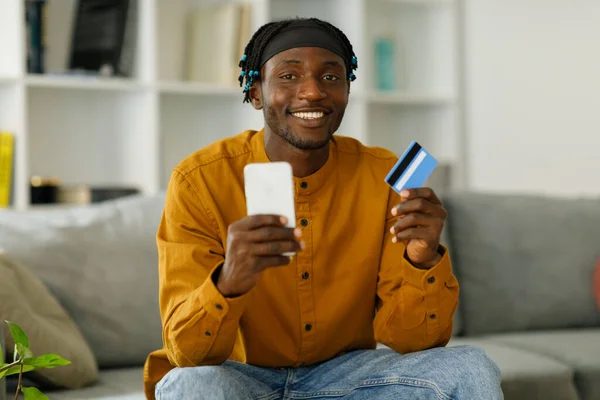 Smiling young black man looking at camera and holding debit credit card using mobile phone, free copy space. High quality photo