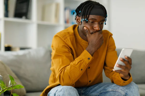 Upset black african man reading bad news in message looking at phone screen sitting on couch. High quality photo