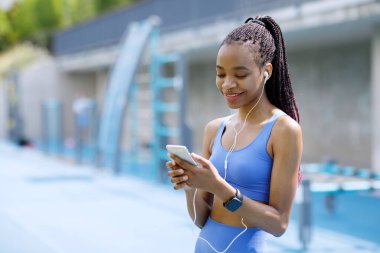 Music and sports. Smiling african american young woman with headphones, smart watch looking at smartphone on sports ground background. High quality photo