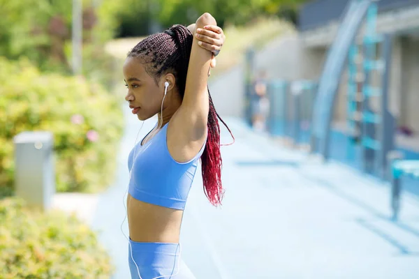 African woman with headphones doing stretching exercises for her arms outdoors. Woman in sportswear doing workout outdoors. High quality photo