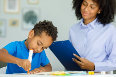 Little boy, training in kindergarten, sitting at the table with a friendly teacher and doing various tests, a young female psychologist working with a preschooler. High quality photo