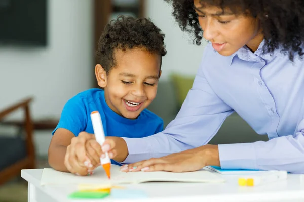 Female infant school teacher working one on one with a young schoolboy, sitting at a table writing in a classroom, front view. High quality photo