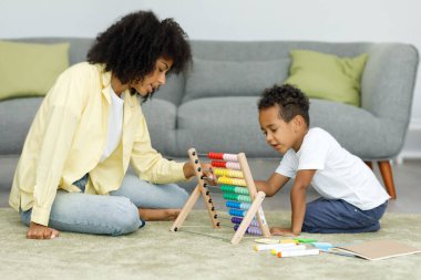 Young african mother with cute little son playing with abacus at home on the floor near the sofa. High quality photo