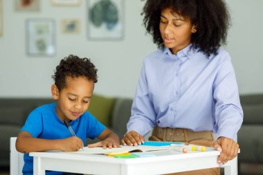 Female infant school teacher working one on one with a young schoolboy, sitting at a table writing in a classroom, front view. High quality photo