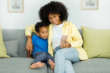 Little ethnic boy watching cartoons on phone smartphone with mother while sitting on couch at home together. High quality photo