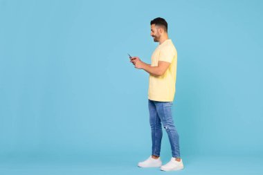 Full body young smiling happy caucasian man holding mobile phone in hand isolated on blue background studio portrait. People lifestyle concept. High quality photo