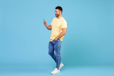 Full body young smiling happy caucasian man holding mobile phone in hand isolated on blue background studio portrait. People lifestyle concept. High quality photo