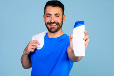 A man in a blue t-shirt and a towel holds a bottle of shampoo or shower gel on a blue background. Shampoo-conditioner for men. Expert care for demanding hair.. High quality photo