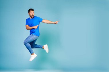 Full length portrait of cheerful young man jumping in blue t-shirt pointing finger to the side celebrating success against blue background. High quality photo