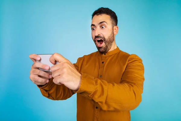 Overjoyed caucasian man in positive shock looks at smartphone on blue studio background. Surprised caucasian man with mobile phone amazed by promo sale or huge discount. High quality photo