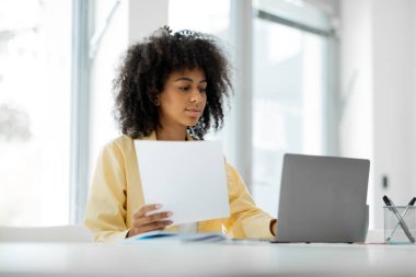A woman is talking on a video connection through a computer, holding a paper in her hands, sitting at a table, working online in a modern office. High quality photo