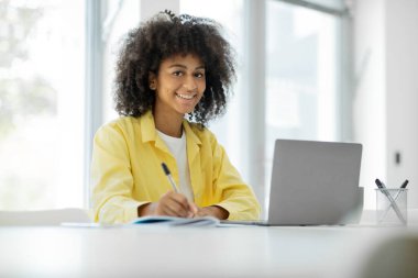 Smiling young black woman looking at camera while sitting at table and working on laptop, free copy space. High quality photo