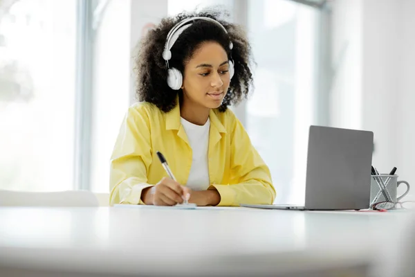 Portrait of a busy focused black woman in a wireless headset writing in a notepad sitting at a work desk with a personal computer in a modern office, profile side view. High quality photo