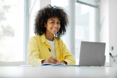 Cheerful black female freelancer looking at camera and working from home sitting at work desk in front of laptop copy space. High quality photo