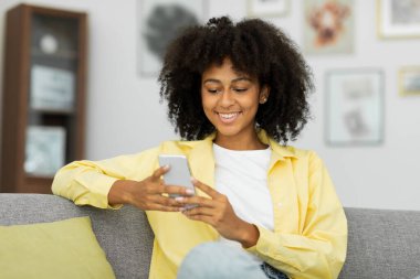 Cheerful black woman using mobile phone typing a message while sitting at the sofa at home. High quality photo
