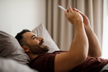 Burgonya pijamalı yakışıklı bir adam cep telefonuyla yatakta uzanıyor, sosyal ağları kontrol ediyor, ev içini kontrol ediyor, fotokopi çekiyor. Yüksek kalite fotoğraf