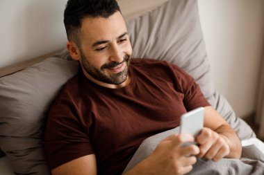 Cep telefonu kullanan, interneti karıştıran, mesajlar gönderen, evde yatakta yatan gülümseyen adam. Yüksek kalite fotoğraf