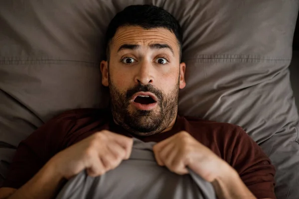 A man in a burgundy T-shirt is scared in bed looking at the camera. High quality photo