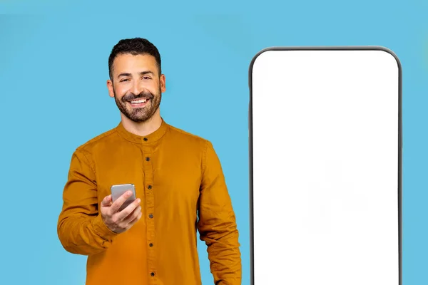 一个穿着黄色衬衫的男人站在一个巨大的智能手机旁边 旁边有一个空白的屏幕 用他的大拇指指向它 复制空间 广告概念 高质量的照片 — 图库照片