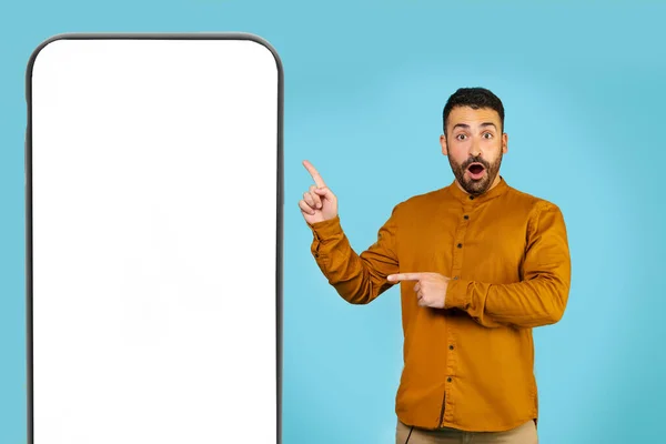 一个穿着黄色衬衫的男人站在一个巨大的智能手机旁边 旁边有一个空白的屏幕 用他的大拇指指向它 复制空间 广告概念 高质量的照片 — 图库照片