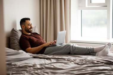 Evde yatakta otururken bilgisayarı serbest çalışan olumlu bir adam. Yüksek kalite fotoğraf