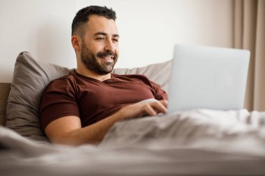 Mutlu sakallı, genç doğulu bir adam yatakta ders çalışıyor, dizüstü bilgisayar kullanıyor, internetten kurs izliyor ya da bir web sitesine katılıyor, kopyalama alanı, yan görüş. Yüksek kalite fotoğraf