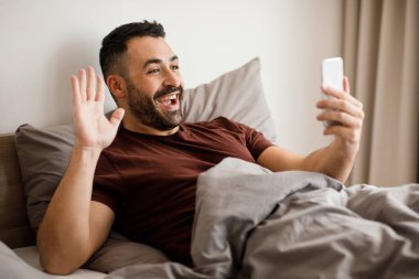 Elinde cep telefonu olan gülümseyen Arap adam video iletişimi kuruyor, evde yatakta yatıyor. Yüksek kalite fotoğraf
