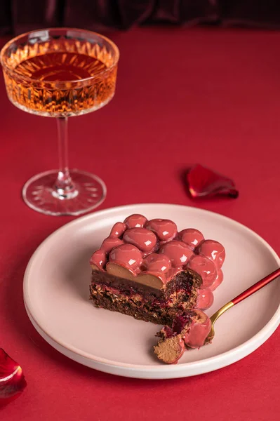 Mousse cake with red mirror icing. Cutaway cake: chocolate sponge cake, crunchy layer, cherry confit, cherry cream, chocolate mousse. Festive cake on a red background with a glass of wine.