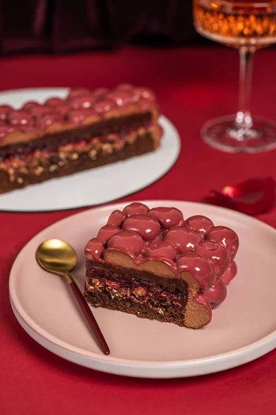 Mousse cake with red mirror icing. Cutaway cake: chocolate sponge cake, crunchy layer, cherry confit, cherry cream, chocolate mousse. Festive cake on a red background with a glass of wine.