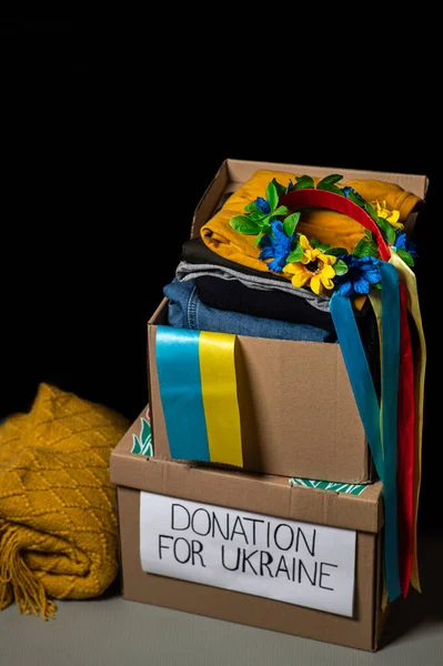 Volunteer packing clothes in donation box with Ukrainian flag. Ukrainian wreath with ribbons for girls. Boxes on a black background.