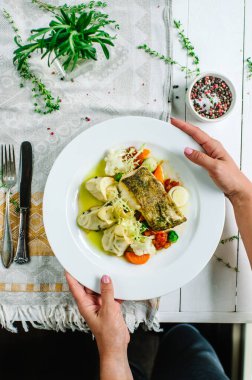 Bitkisel yağlı beyaz balık. Zeytinli patates püresi ve karışık sebzeler - karnabahar, havuç, domates ve Brüksel lahanası. Bir kadın elinde bir tabak tutuyor. Fotoğraf kırsal stilde.
