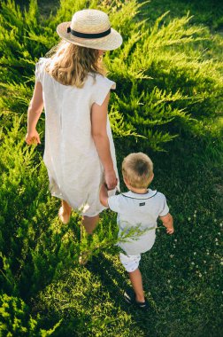 Anne oğluyla birlikte doğada yürüyor. Arkadan üstten bak. Anne küçük oğlunun elini tutuyor. Yeşil çimlerin üzerinde yürü.
