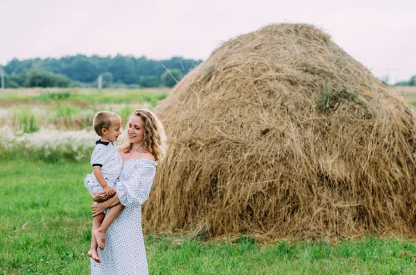 Happy family. A young woman with blond curly hair holds her son in her arms against the background of hay. Mother and son. Summer mood
