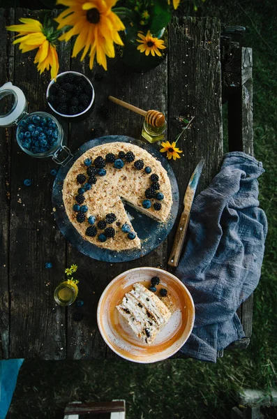 Homemade honey cake with prunes on a wooden table in nature. Cake decorated with blueberries and blackberries. Summer cake. Top view. Photo in rustic style.