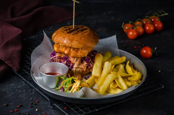 Fresh and tasty crispy chicken burger with melted cheese, red cabbage and sauce on a metal plate. Served with french fries, salad and ketchup. Dish in a restaurant.