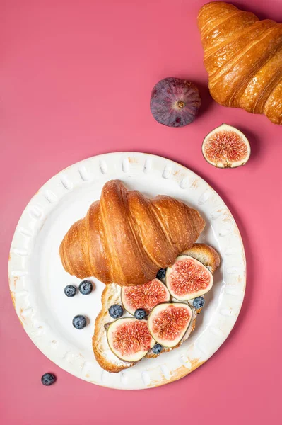 Modern photo on a bright pink background. Croissants with cream cheese, figs, blueberries and honey on a light plate. Breakfast concept, top view. Croissant with healthy dietary filling.