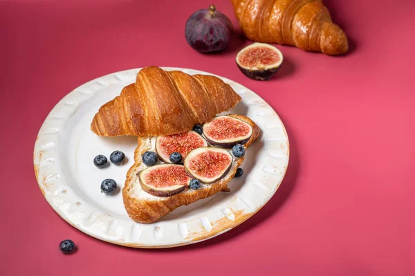 Modern photo on a bright pink background. Croissants with cream cheese, figs, blueberries and honey on a light plate. Breakfast concept, top view. Croissant with healthy dietary filling.