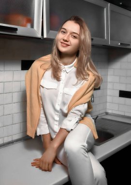 Beyaz gömlekli, pantolonlu, mutfak masasında poz veren güzel bir genç kadın.