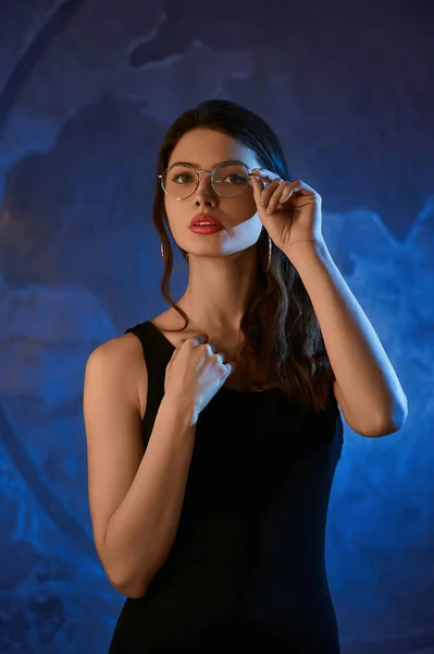 Beautiful young Girl in image of geography teacher in blue neon light in transparent round glasses in black dress looks at camera while standing against background of world atlas. 
