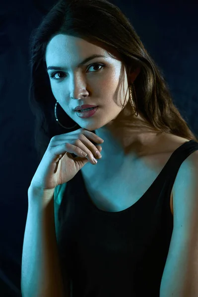 Portrait of beautiful young brunette girl looking at camera, propping her chin with her hand in light of cyan neon light on black background.