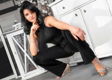 Siyah bluzlu ve pantolonlu genç esmer kadın topuklularla çömeliyor, çenesini parmağıyla destekliyor, beyaz çekmecelerin arka planında kameraya bakıyor..