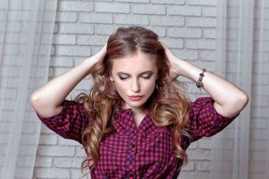 Beautiful young girl with long curls of hair with her eyes closed holds her hands behind her head against background of white brick wall.